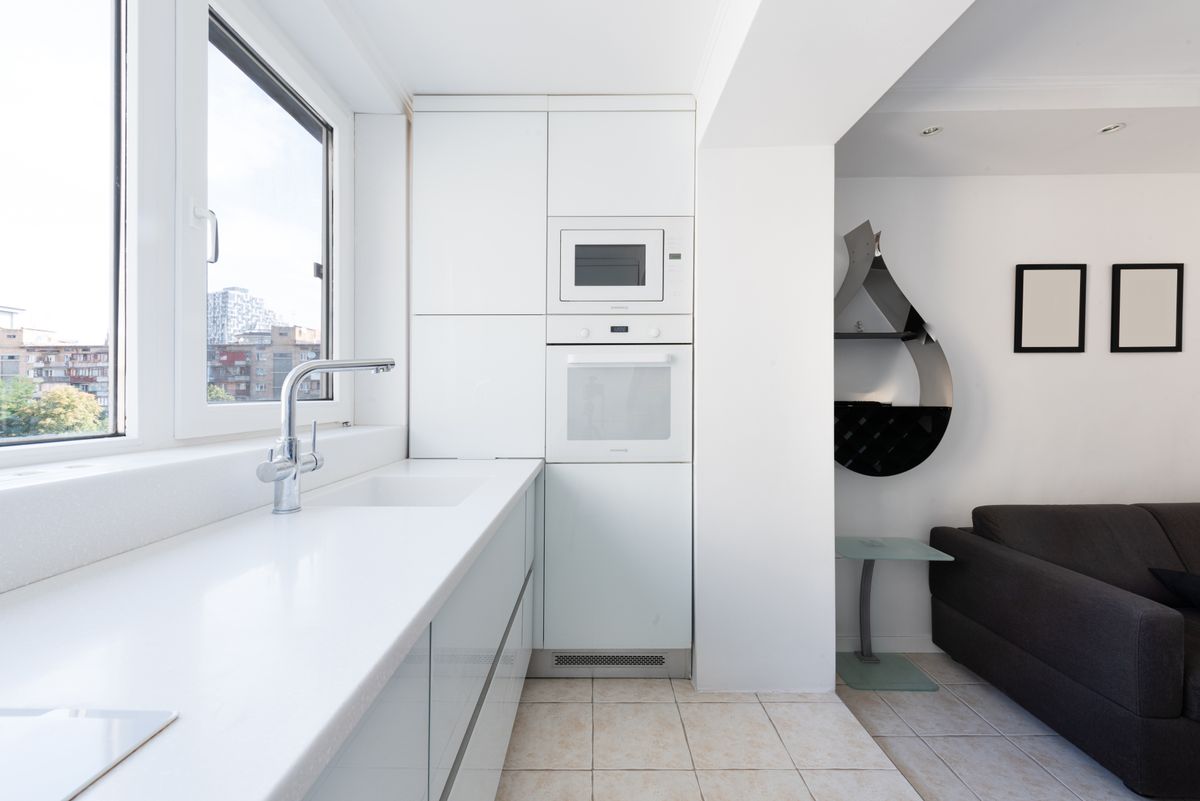 Modern studio interior with kitchen cabinets with sink and tap near oven and microwave near windows and living room with sofa near table and shelves on wall in daylight